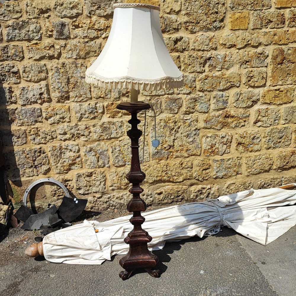A dark carved wooden standard lamp featuring a baluster column and topped with a cream fabric lampshade trimmed with fringe.