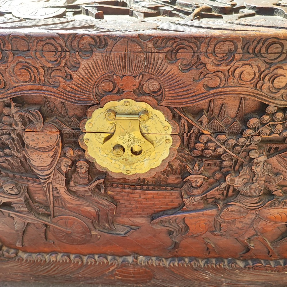 A relief-carved camphor wood trunk with relief panel depictions of figures on horseback and a central brass clasp. View II