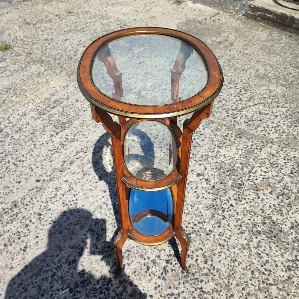 An antique oval satinwood etagere featuring three tiers with glass panels, brass trim, and curved legs adorned with bronze mounts. View III