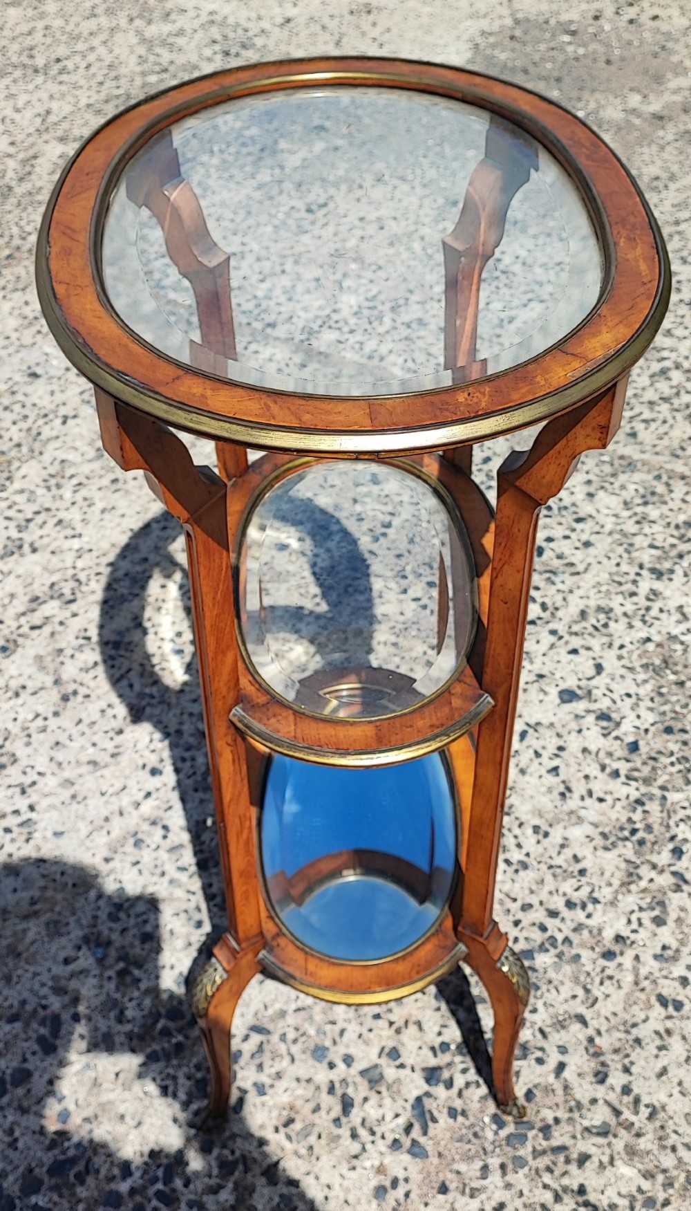 An antique oval satinwood etagere featuring three tiers with glass panels, brass trim, and curved legs adorned with bronze mounts. View VII