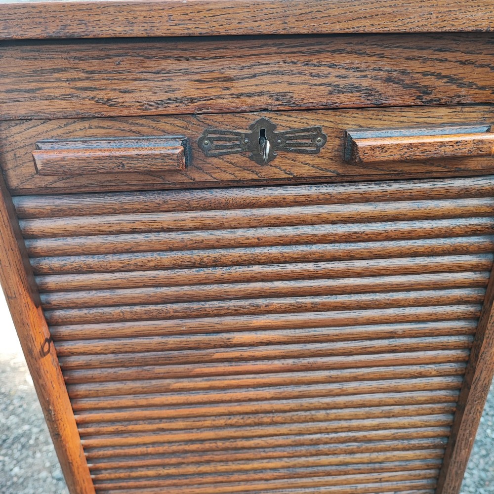 A tall Edwardian oak filing cabinet featuring a slatted wooden tambour front shutter, keyhole escutcheon with key, and wooden pull handles. View VII