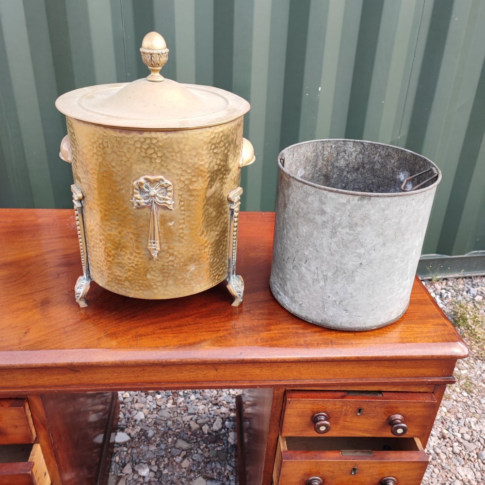 A cylindrical hammered brass coal bin with an acorn finial lid, ribbon bow decoration, paw feet, and a removable galvanised metal liner.