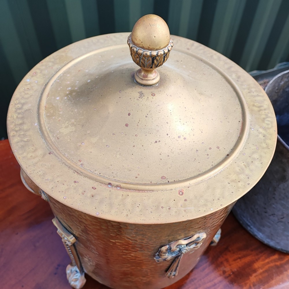 A cylindrical hammered brass coal bin with an acorn finial lid, ribbon bow decoration, paw feet, and a removable galvanised metal liner. View II