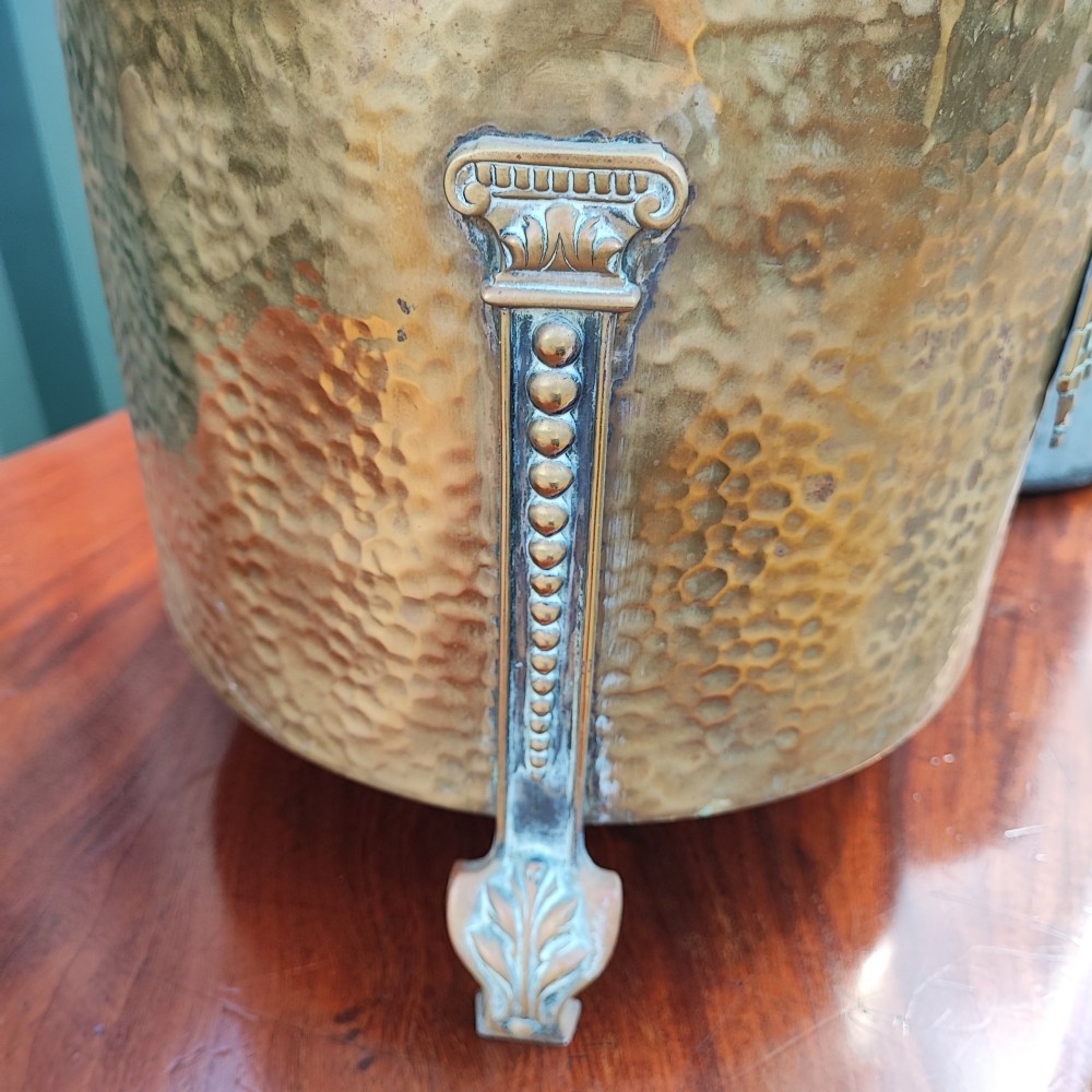A cylindrical hammered brass coal bin with an acorn finial lid, ribbon bow decoration, paw feet, and a removable galvanised metal liner. View V