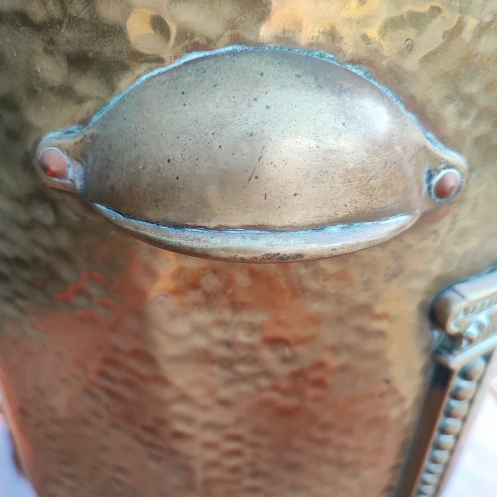 A cylindrical hammered brass coal bin with an acorn finial lid, ribbon bow decoration, paw feet, and a removable galvanised metal liner. View VI