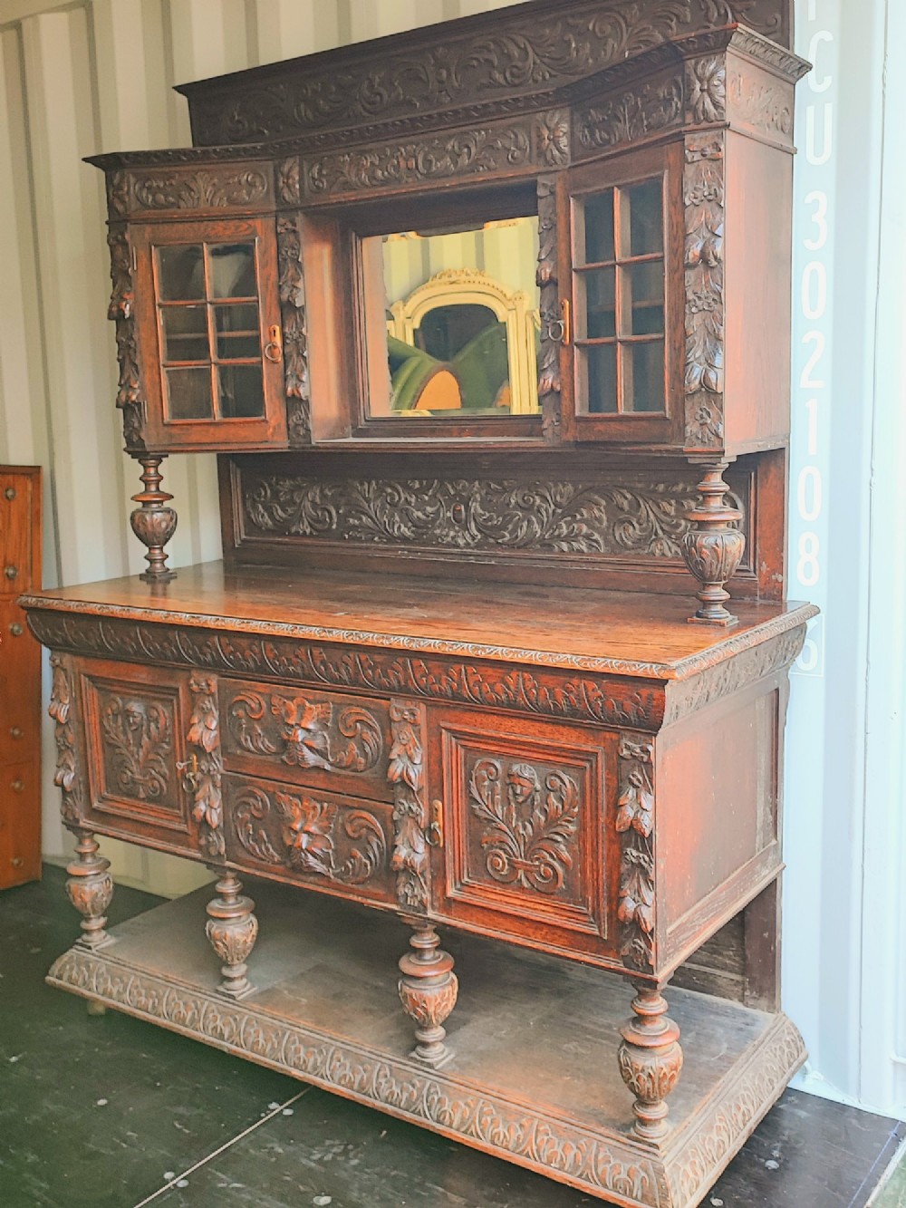 A heavily carved dark oak sideboard featuring a mirrored top section flanked by glazed cabinet doors, resting on turned bulbous supports and a lower shelf.