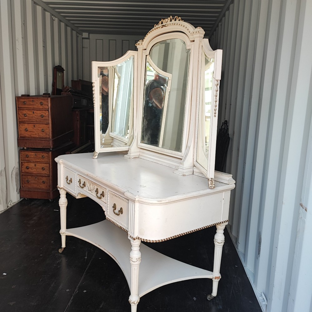 An off-white painted antique dressing table featuring a central triptych mirror, carved gilt details, three drawers with brass handles, and fluted legs on castors. View II