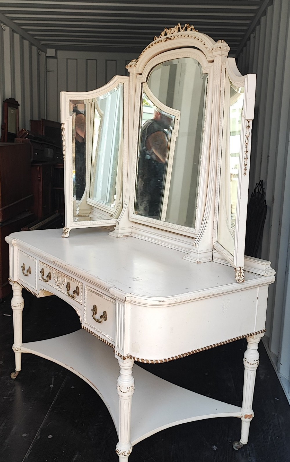 An off-white painted antique dressing table featuring a central triptych mirror, carved gilt details, three drawers with brass handles, and fluted legs on castors. View V