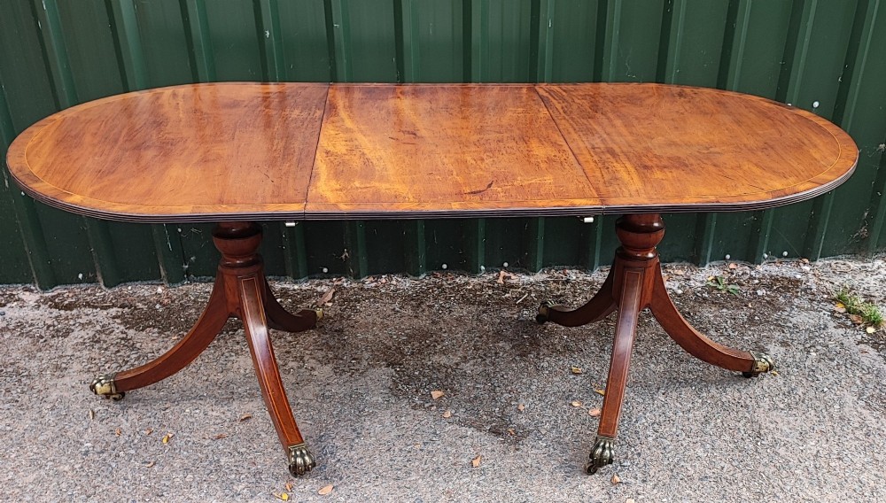 A warm mahogany twin pedestal dining table featuring an oval crossbanded top with string inlay and splayed legs resting on brass claw castors.