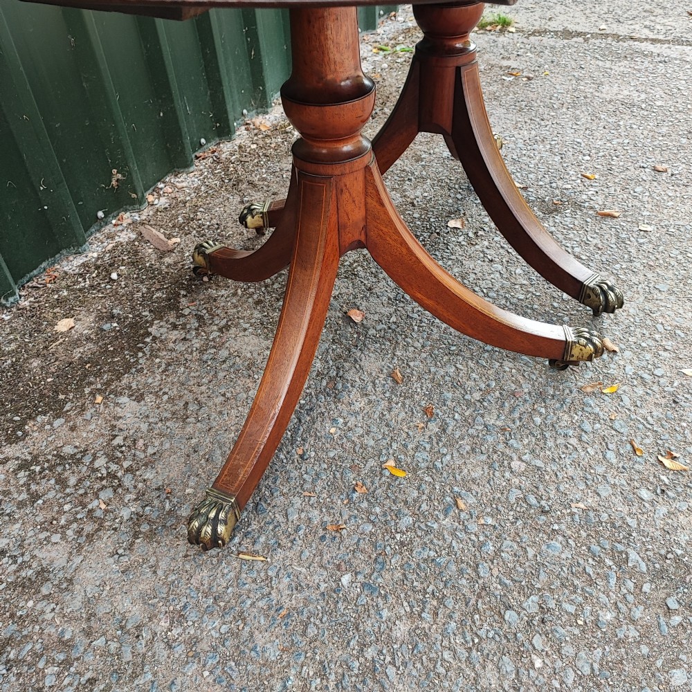A warm mahogany twin pedestal dining table featuring an oval crossbanded top with string inlay and splayed legs resting on brass claw castors. View II