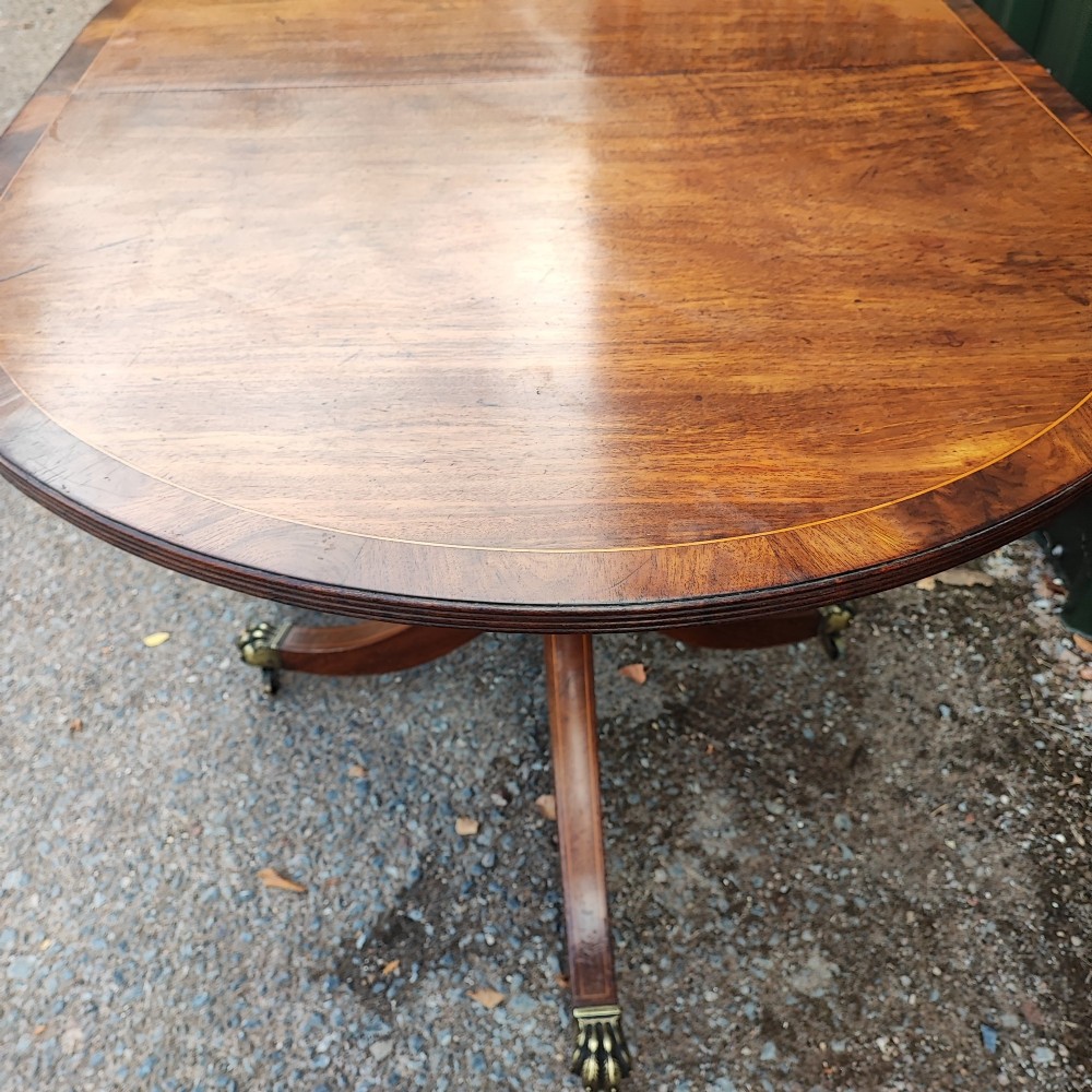 A warm mahogany twin pedestal dining table featuring an oval crossbanded top with string inlay and splayed legs resting on brass claw castors. View V