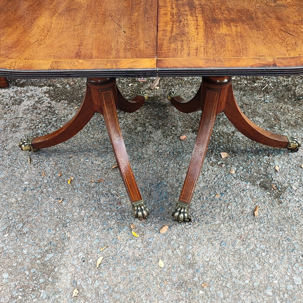 A warm mahogany twin pedestal dining table featuring an oval crossbanded top with string inlay and splayed legs resting on brass claw castors. View VI