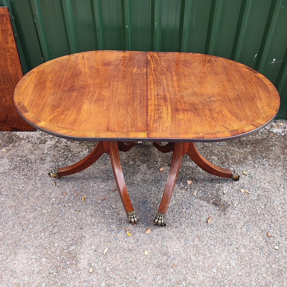 A warm mahogany twin pedestal dining table featuring an oval crossbanded top with string inlay and splayed legs resting on brass claw castors. View VII