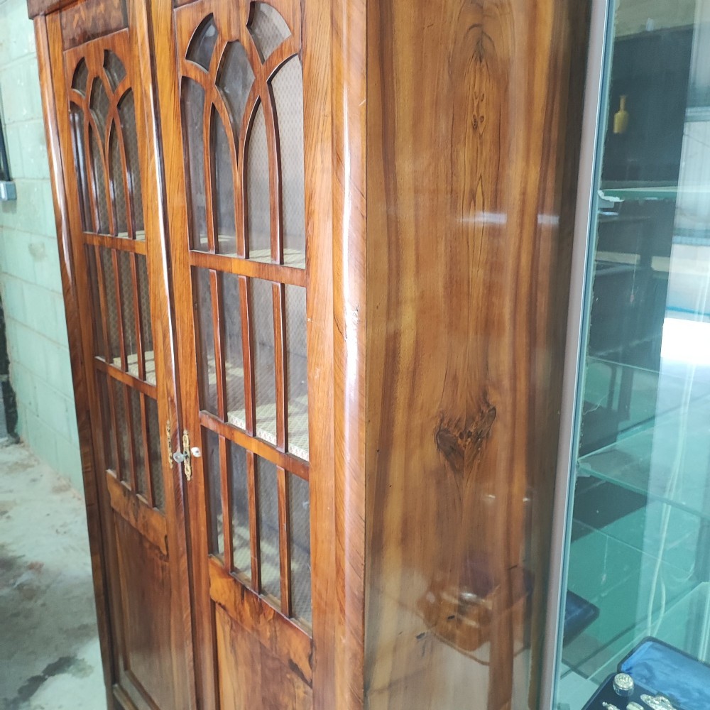A tall 19th-century walnut display cabinet featuring two glazed doors with Gothic arched tracery above solid figured wood lower panels. View II