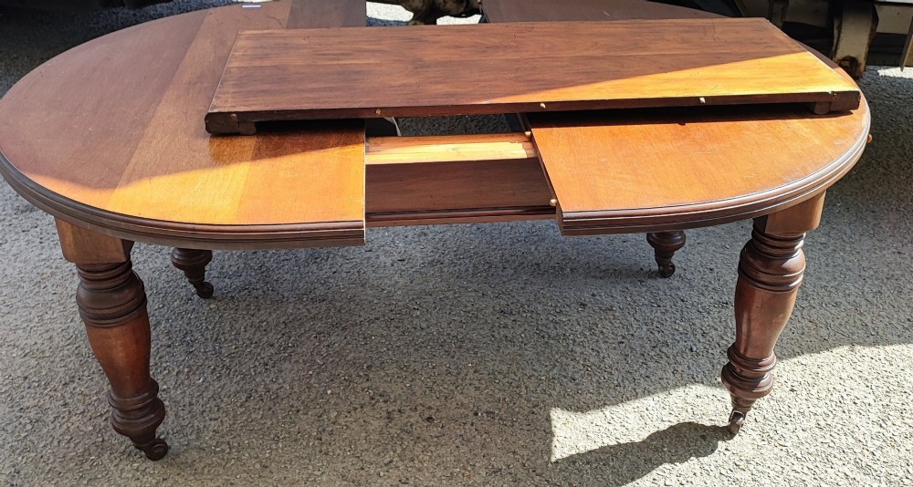 A circular Victorian mahogany dining table with substantial turned legs on castors, featuring a loose extension leaf and winding handle resting on top. View II