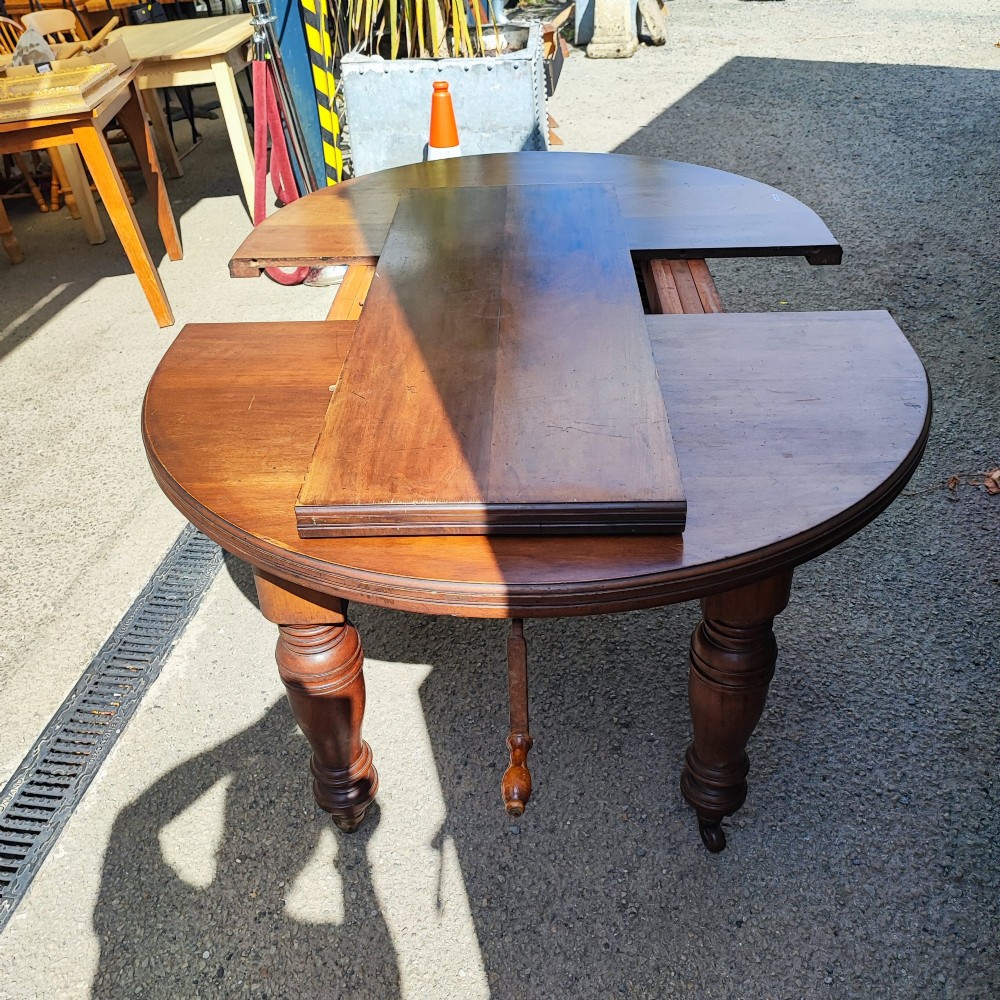 A circular Victorian mahogany dining table with substantial turned legs on castors, featuring a loose extension leaf and winding handle resting on top. View VI