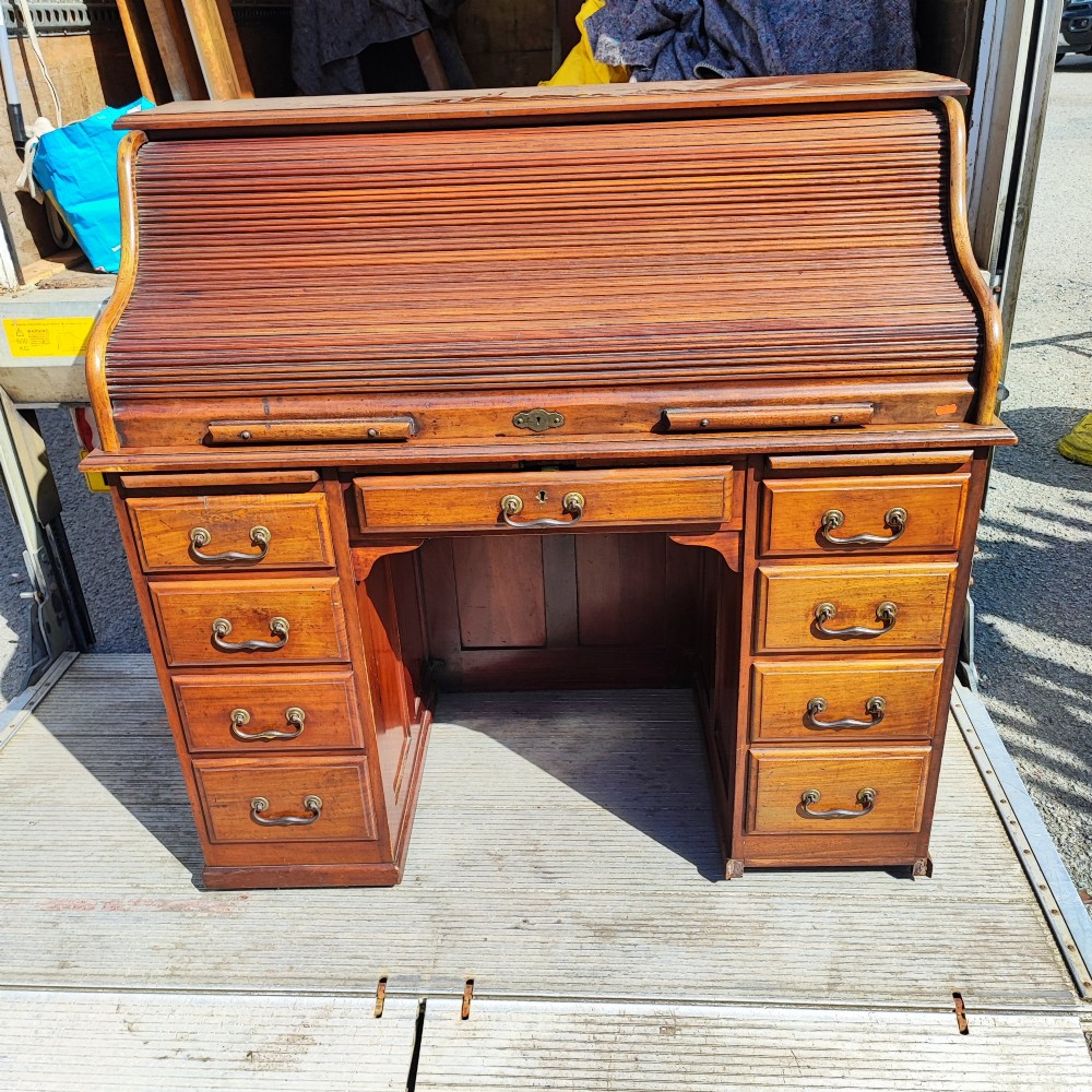 An antique mahogany roll top desk featuring a curved tambour front shutter, twin pedestals with multiple drawers, and brass pull handles. View VI