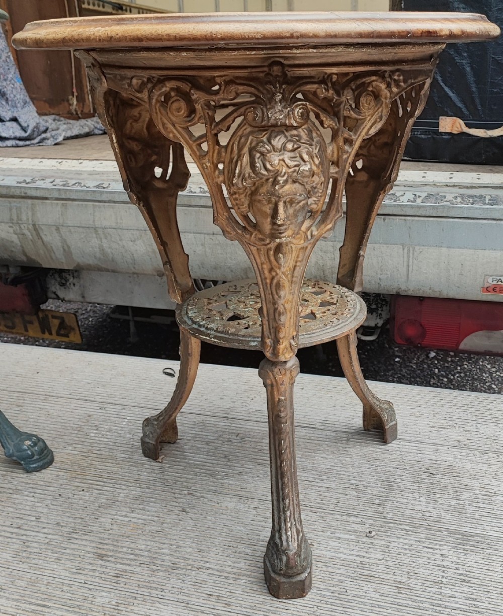 A Victorian cast iron tripod pub table featuring ornate leg castings with classical face motifs, a pierced lower circular tier, and a round wooden tabletop.