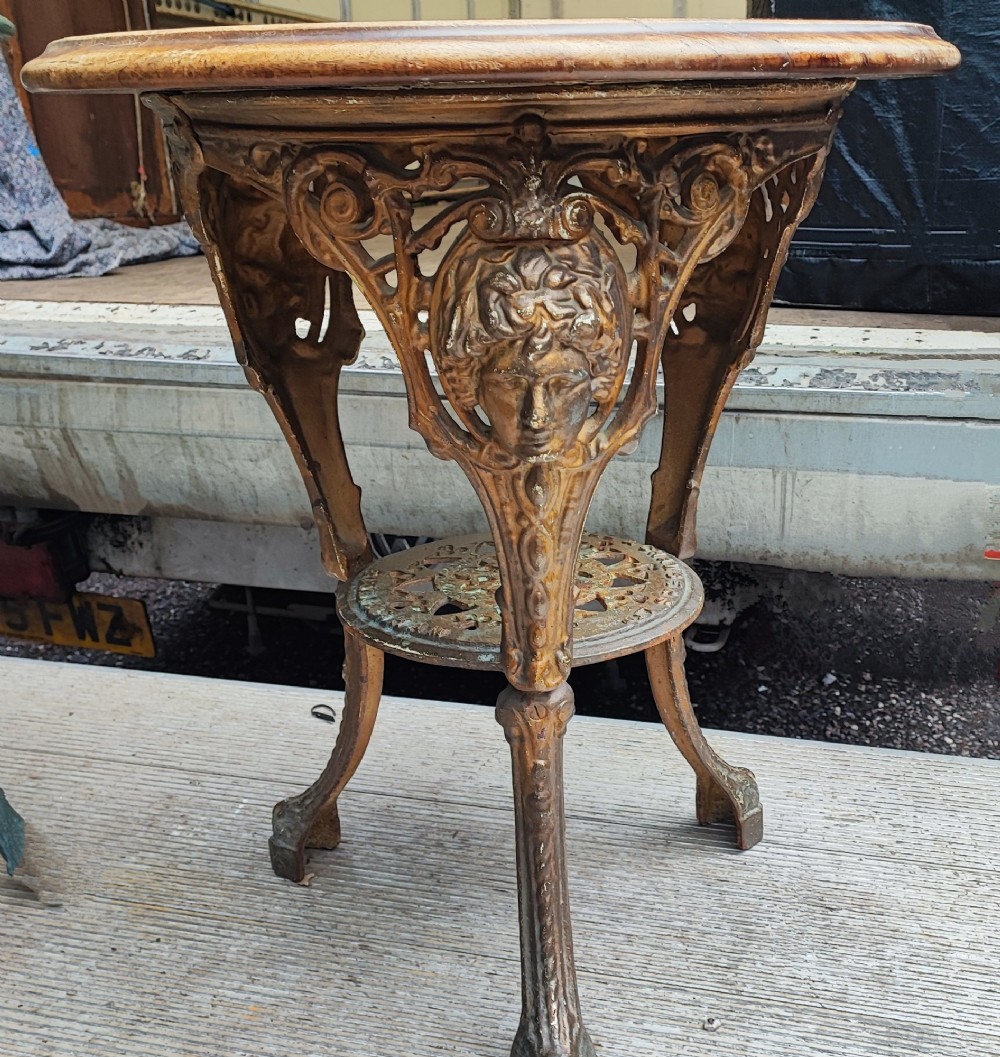 A Victorian cast iron tripod pub table featuring ornate leg castings with classical face motifs, a pierced lower circular tier, and a round wooden tabletop. View V