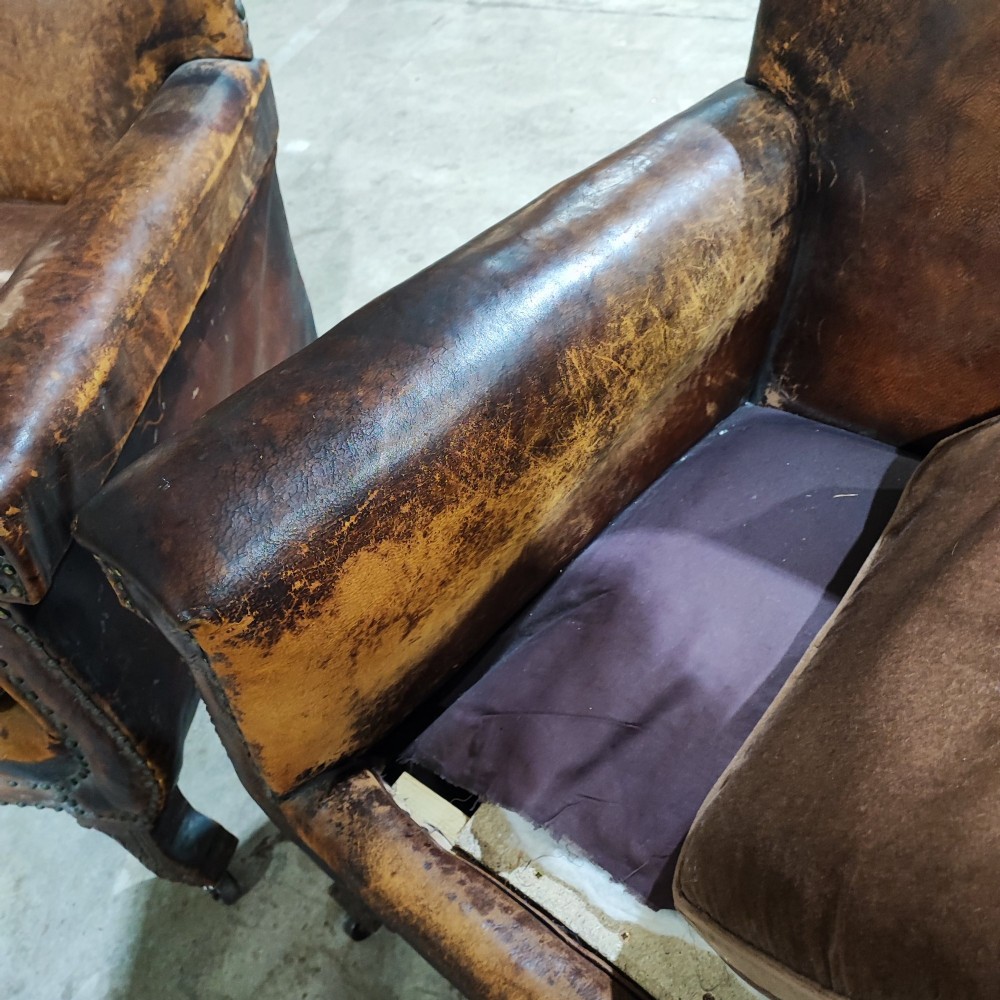 A pair of patinated tan brown leather club armchairs featuring shaped backrests, brass studding along the trim, brown velvet seat cushions, and short carved front legs on brass castors. View V