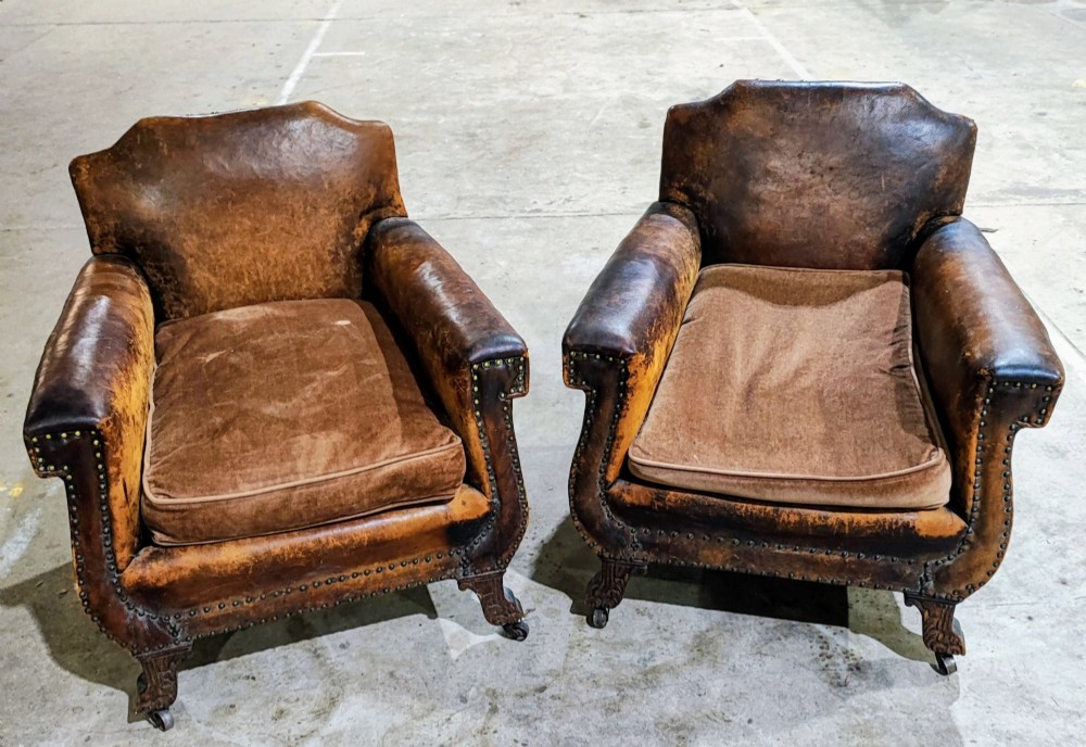 A pair of patinated tan brown leather club armchairs featuring shaped backrests, brass studding along the trim, brown velvet seat cushions, and short carved front legs on brass castors. View VI