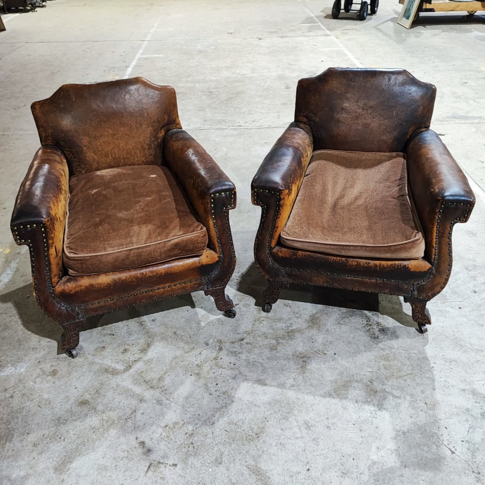 A pair of patinated tan brown leather club armchairs featuring shaped backrests, brass studding along the trim, brown velvet seat cushions, and short carved front legs on brass castors. View VII