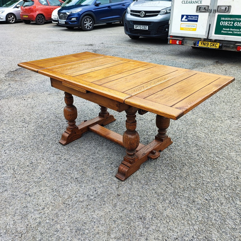 A solid oak refectory dining table featuring a draw-leaf extension mechanism, carved apron, turned baluster legs, and a low central stretcher base. View V