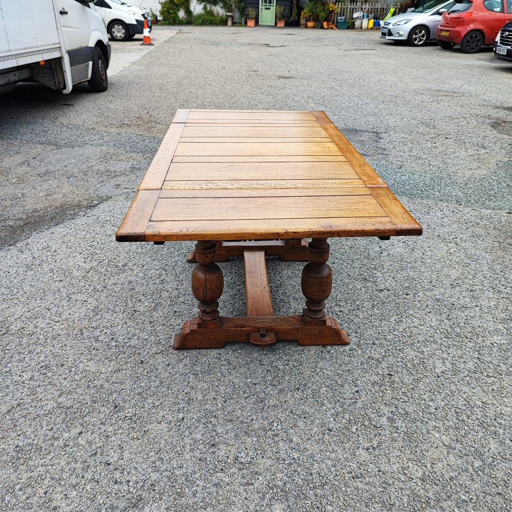 A solid oak refectory dining table featuring a draw-leaf extension mechanism, carved apron, turned baluster legs, and a low central stretcher base. View VII