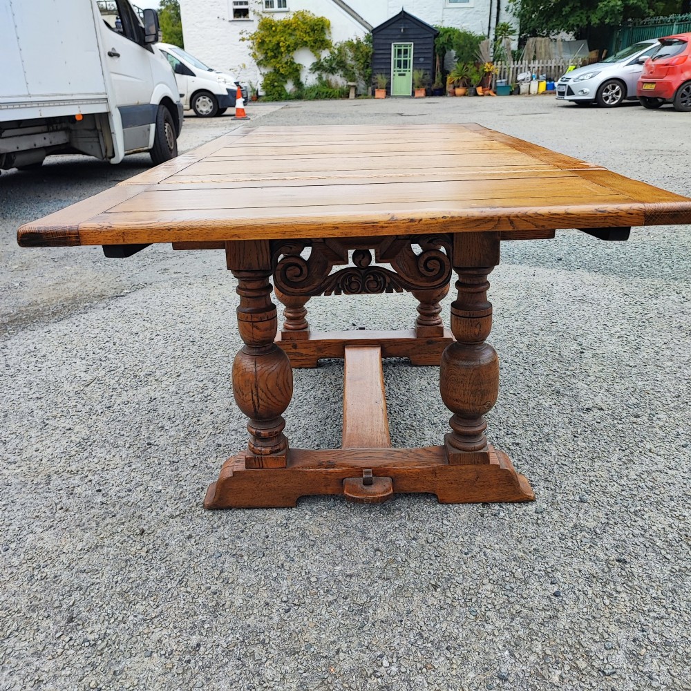 A solid oak refectory dining table featuring a draw-leaf extension mechanism, carved apron, turned baluster legs, and a low central stretcher base. View VIII