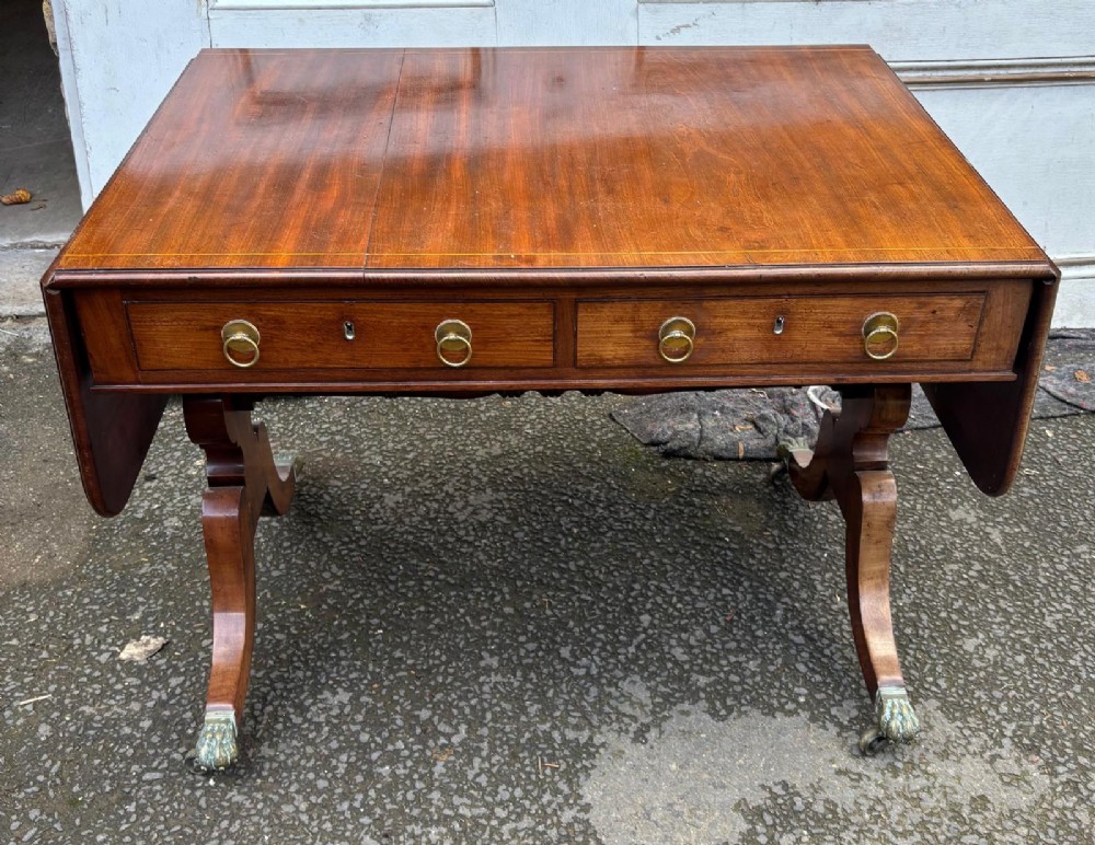 A Regency mahogany sofa table featuring two hinged drop leaves, two frieze drawers with brass ring handles, and splayed legs ending in brass paw castors.