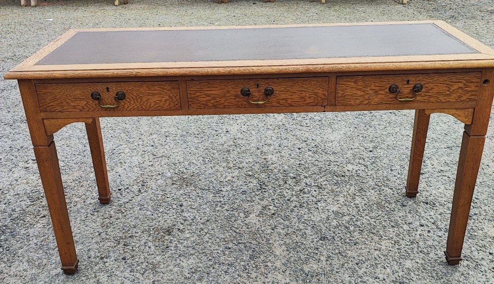A medium brown oak writing table with three inline drawers, brass drop handles, a leather insert top, and four square tapered legs. View II