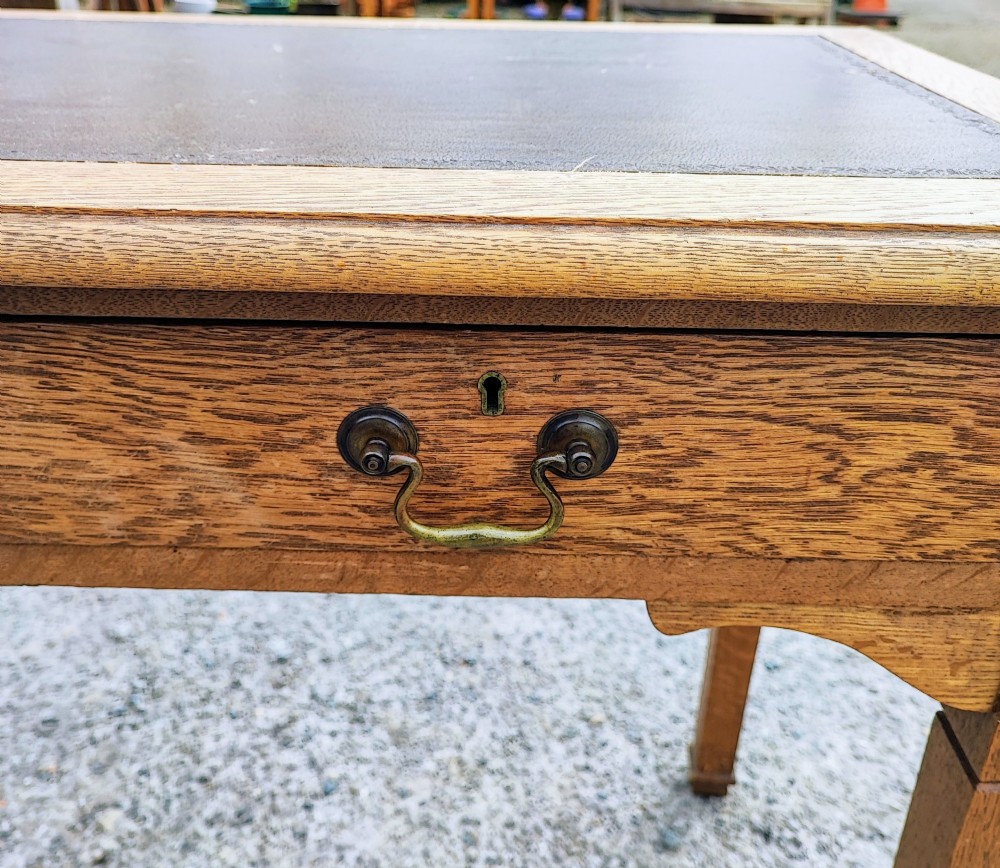 A medium brown oak writing table with three inline drawers, brass drop handles, a leather insert top, and four square tapered legs. View III