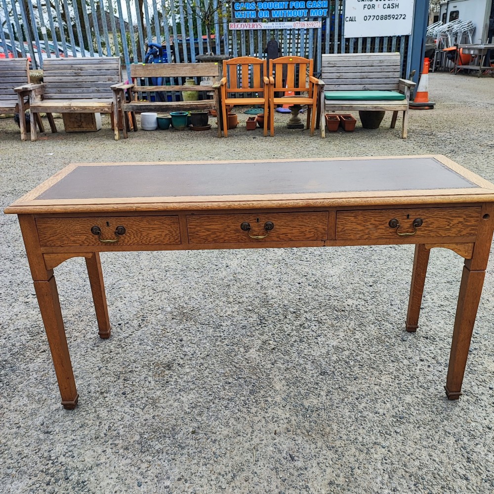 A medium brown oak writing table with three inline drawers, brass drop handles, a leather insert top, and four square tapered legs. View IV