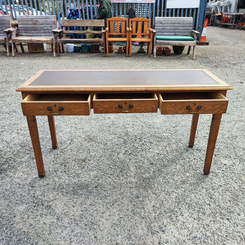 A medium brown oak writing table with three inline drawers, brass drop handles, a leather insert top, and four square tapered legs. View V