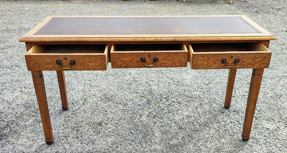 A medium brown oak writing table with three inline drawers, brass drop handles, a leather insert top, and four square tapered legs. View VI