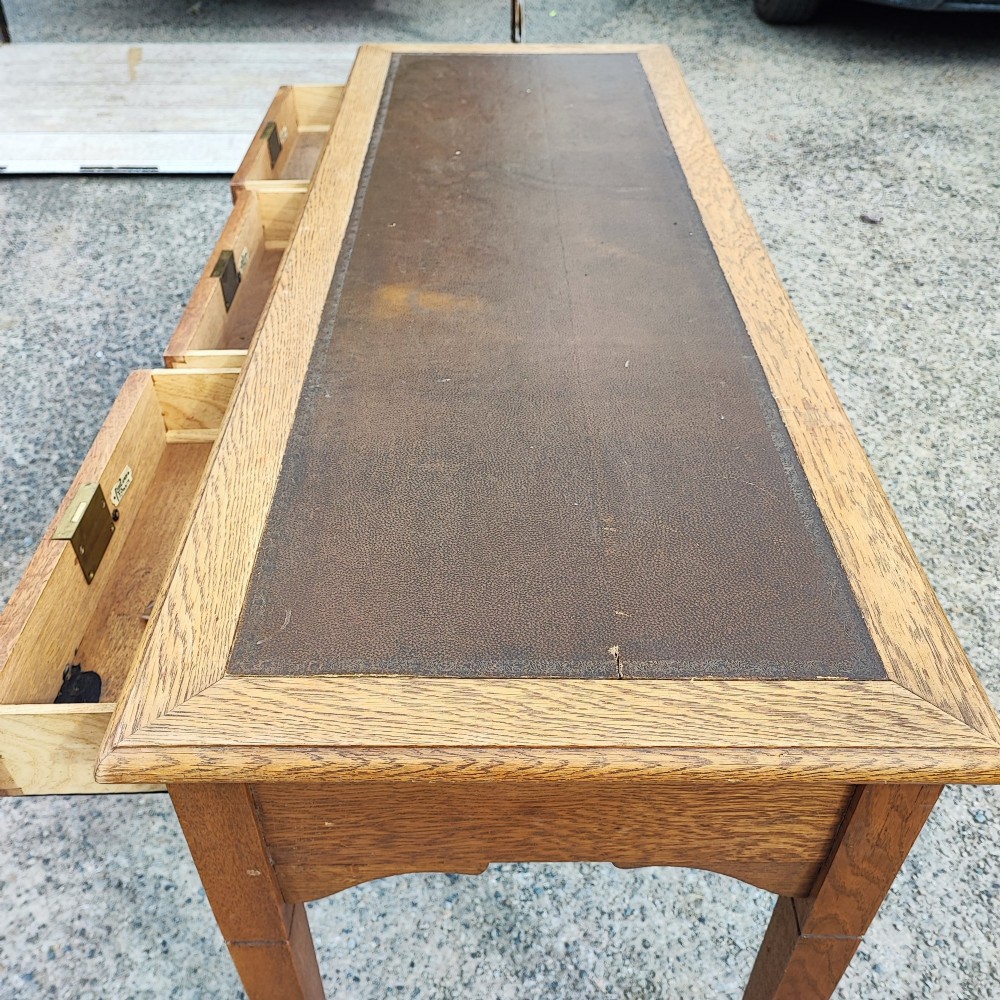 A medium brown oak writing table with three inline drawers, brass drop handles, a leather insert top, and four square tapered legs. View VIII