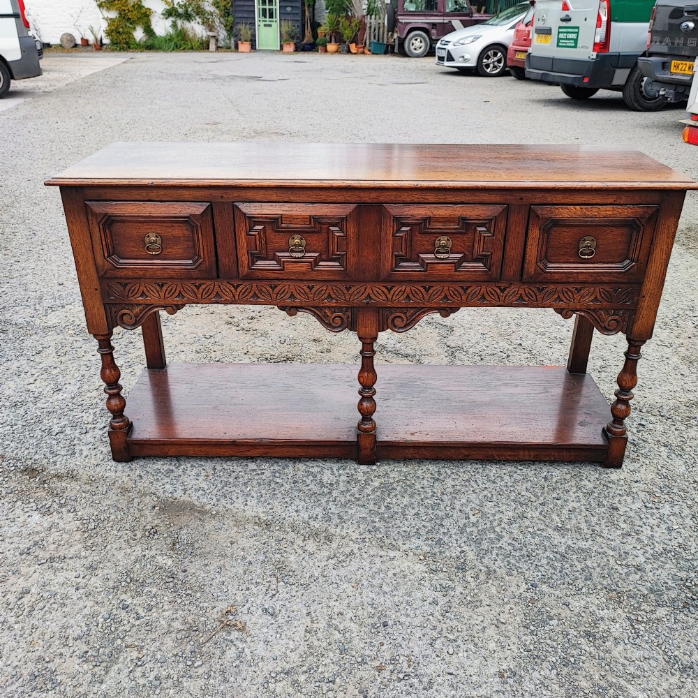 A low carved oak dresser base featuring geometric drawer mouldings, turned front legs, brass ring pulls, and an open lower pot shelf. View II