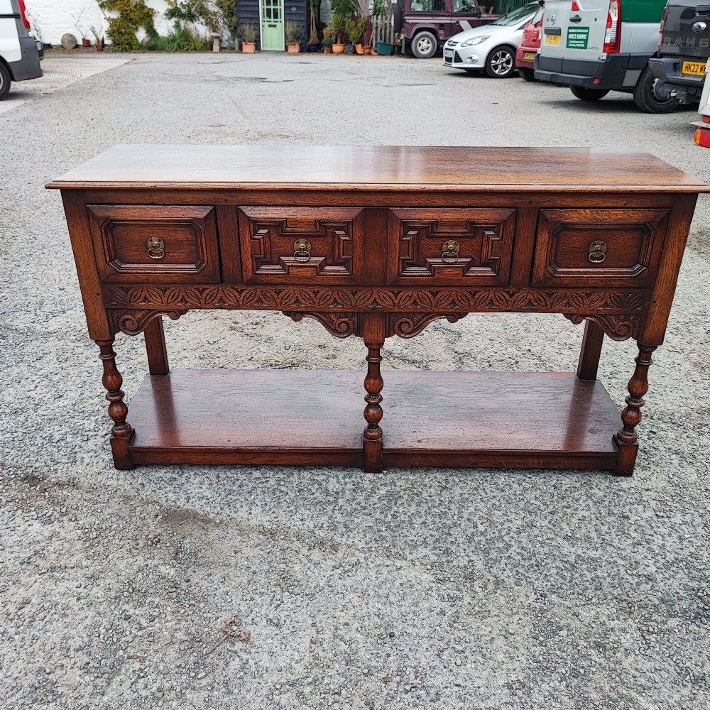 A low carved oak dresser base featuring geometric drawer mouldings, turned front legs, brass ring pulls, and an open lower pot shelf. View VII