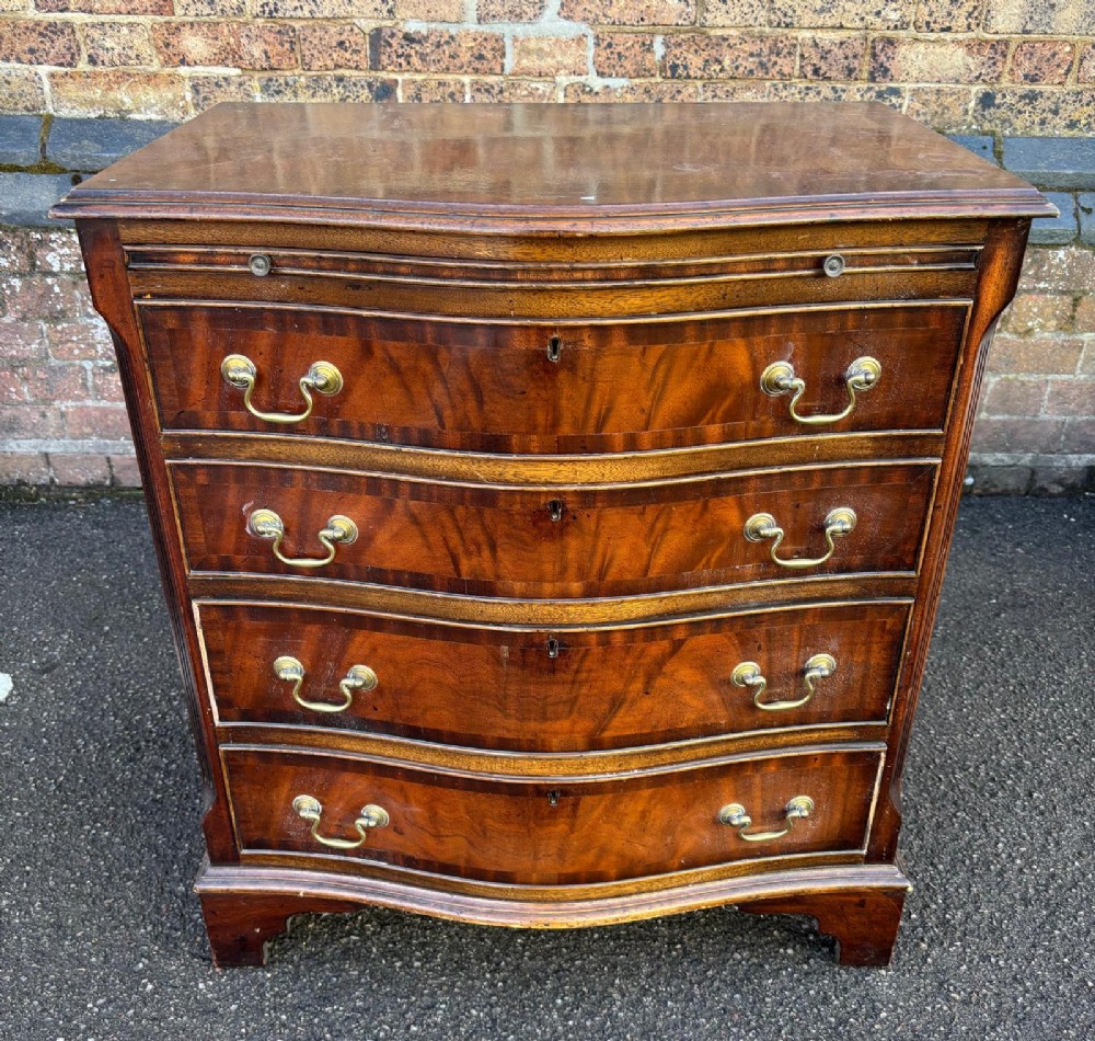 An antique mahogany chest of drawers featuring a serpentine front, four stacked drawers with brass handles, a pull-out brushing slide, and bracket feet.