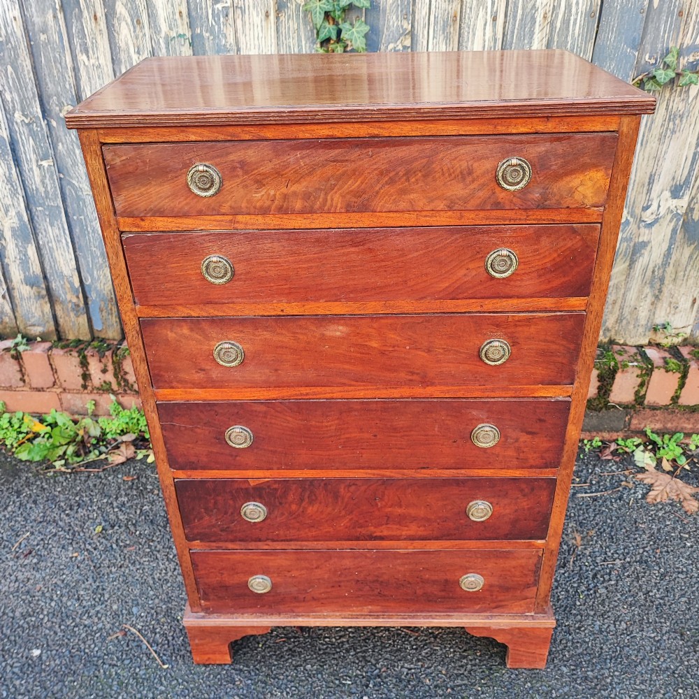 A tall, narrow mahogany chest of six drawers with circular brass ring pulls, flame veneer drawer fronts, and bracket feet. View III
