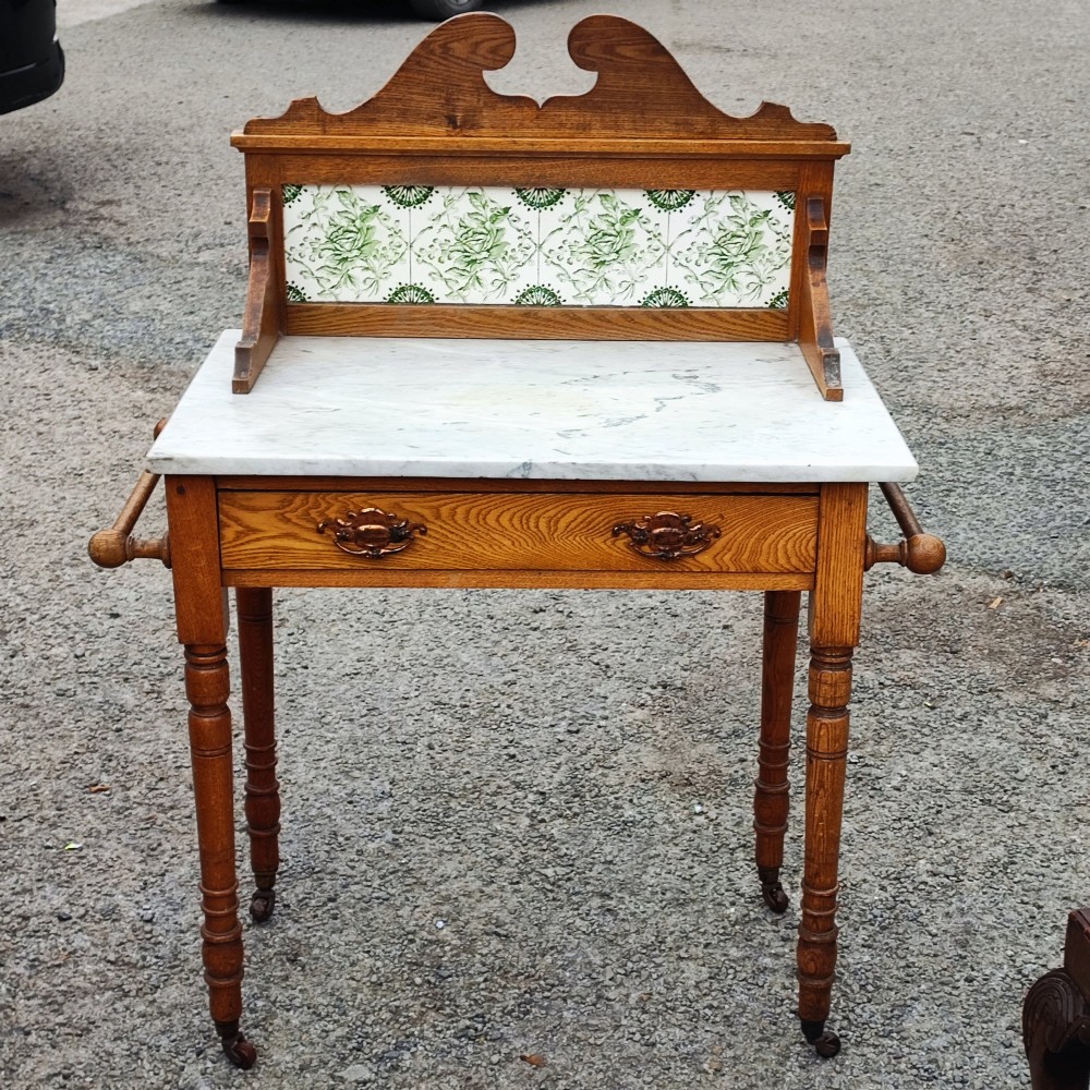 An Arts and Crafts ash timber washstand featuring a white marble top, green patterned tiled splashback, side towel rails, and turned legs.