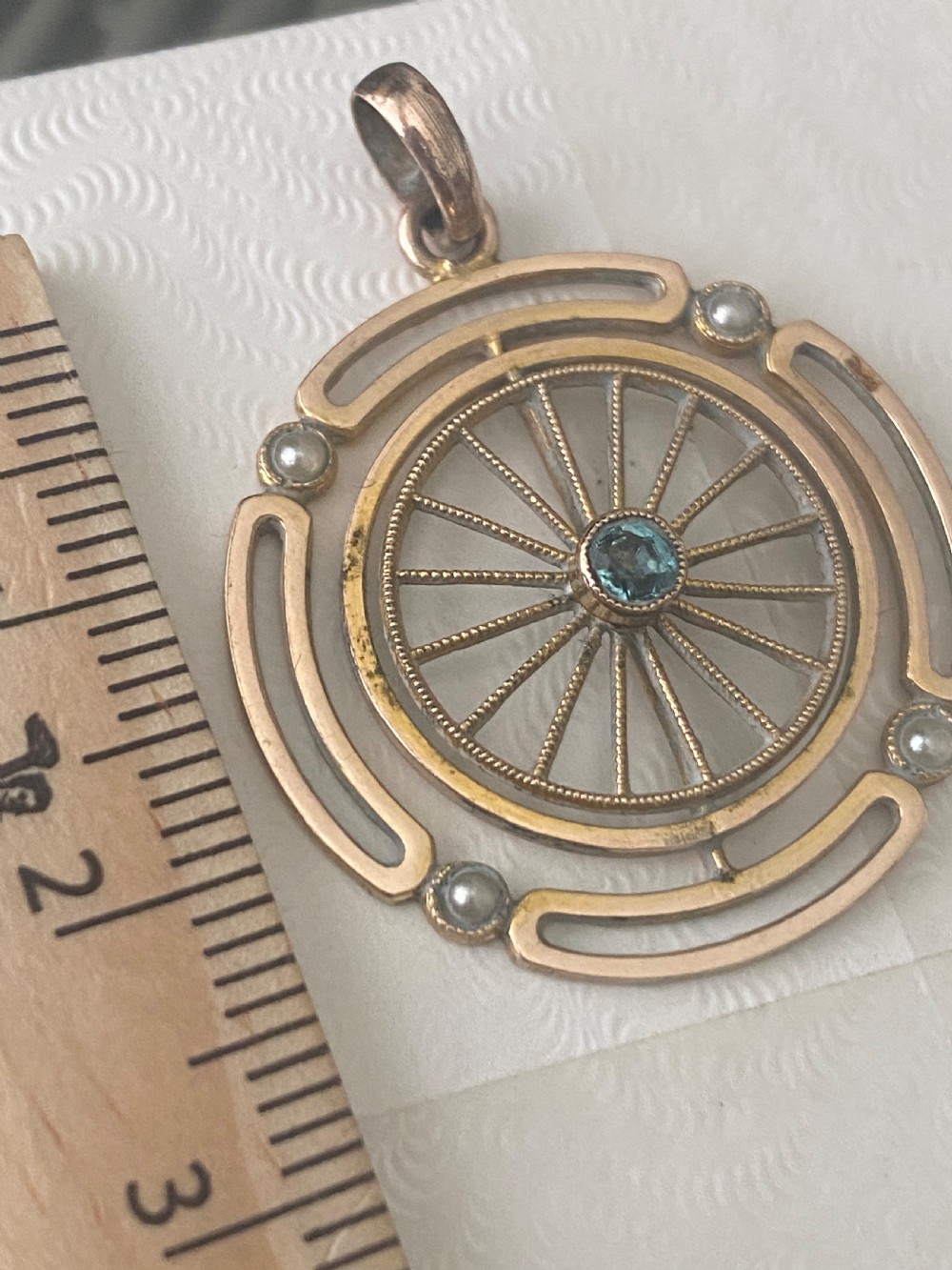 A circular Edwardian yellow gold pendant featuring an openwork spoked wheel design with a blue gemstone at the centre and pearl accents on the outer frame. View VI