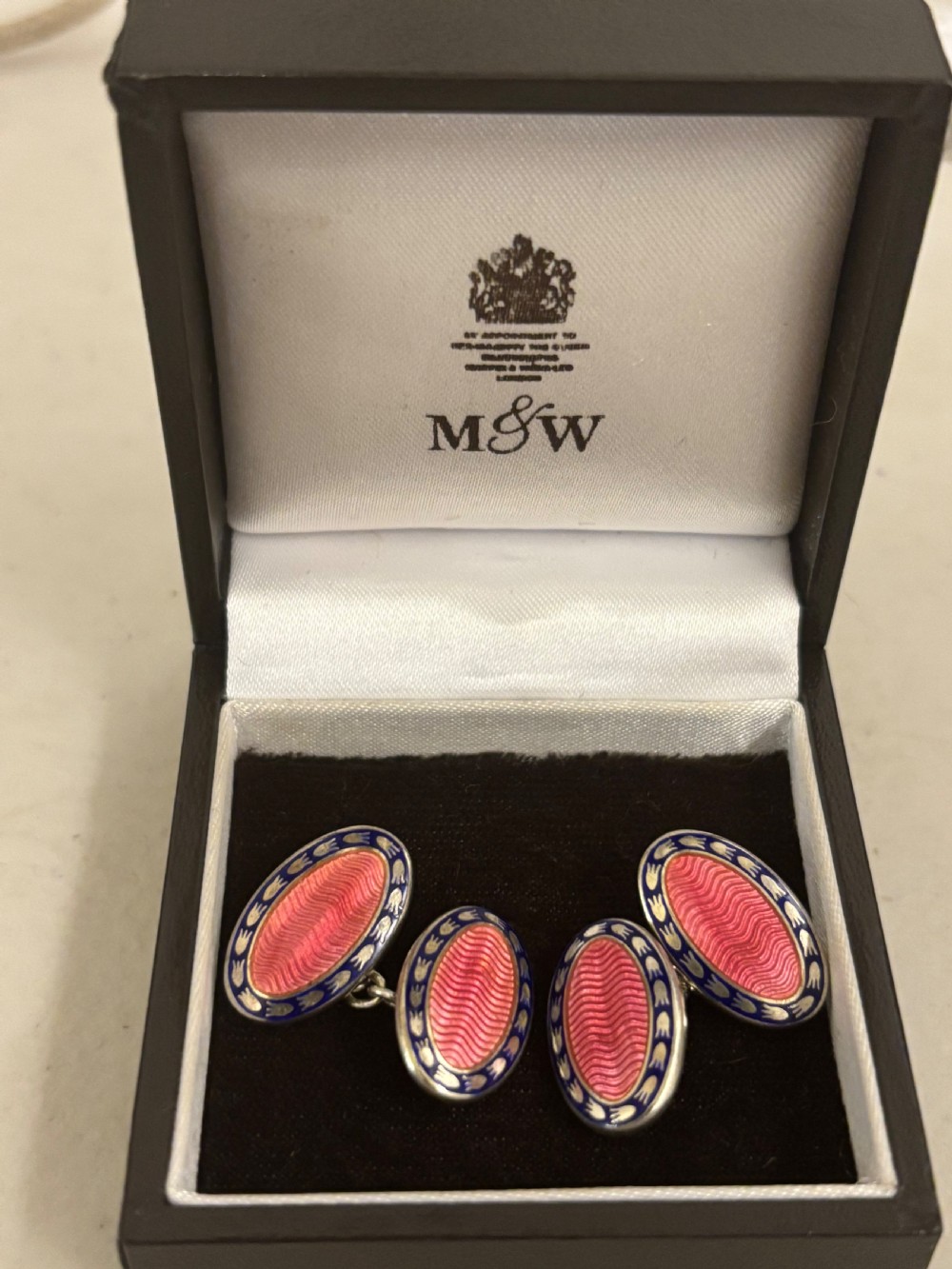 A pair of oval silver cufflinks featuring pink guilloche enamel centres surrounded by blue and white enamel borders, resting inside a branded presentation box.