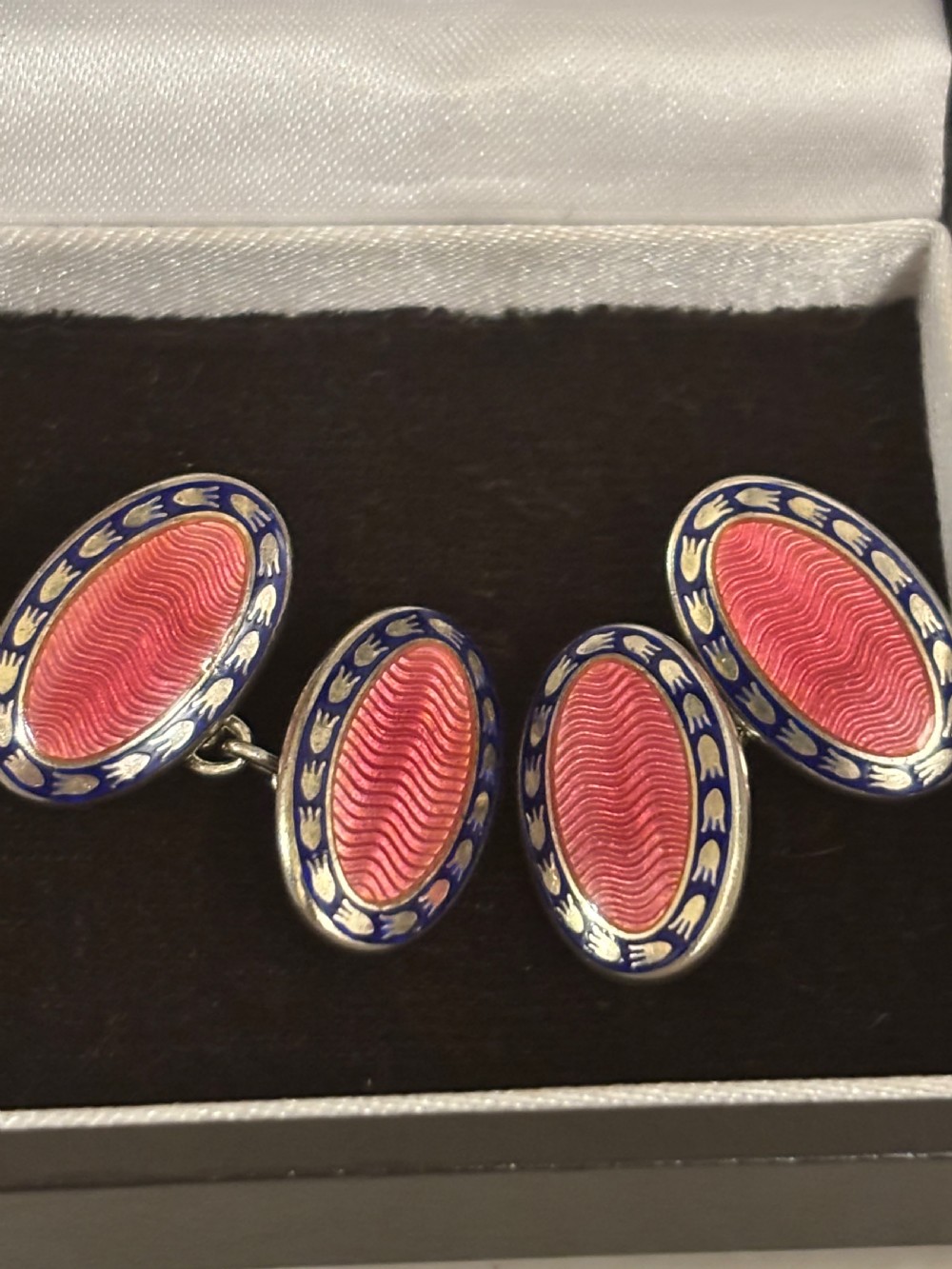 A pair of oval silver cufflinks featuring pink guilloche enamel centres surrounded by blue and white enamel borders, resting inside a branded presentation box. View II