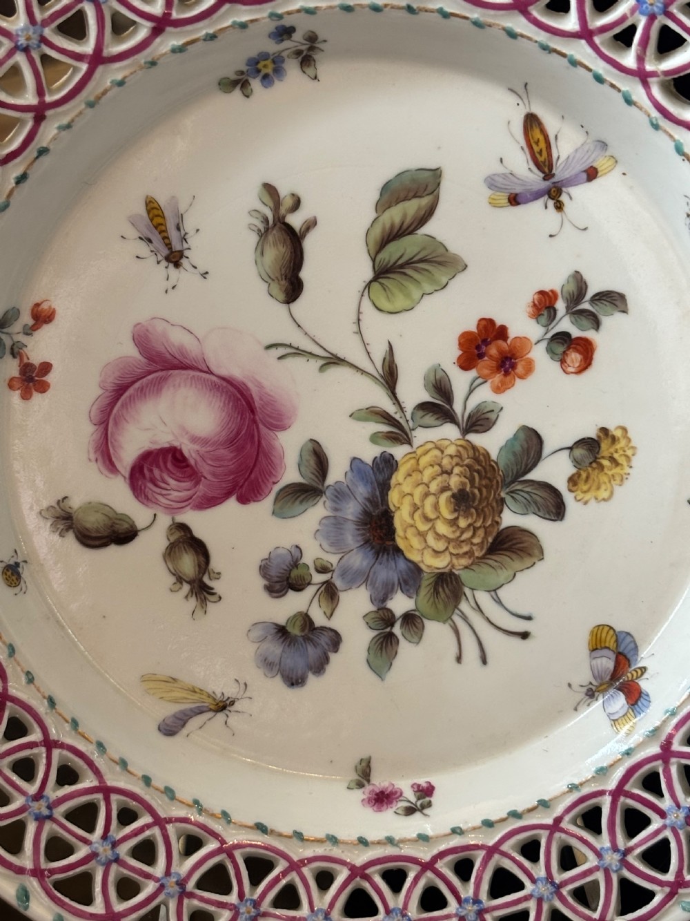 A circular porcelain plate featuring a pink reticulated lattice rim and a hand-painted central botanical scene with flowers and insects. View XI