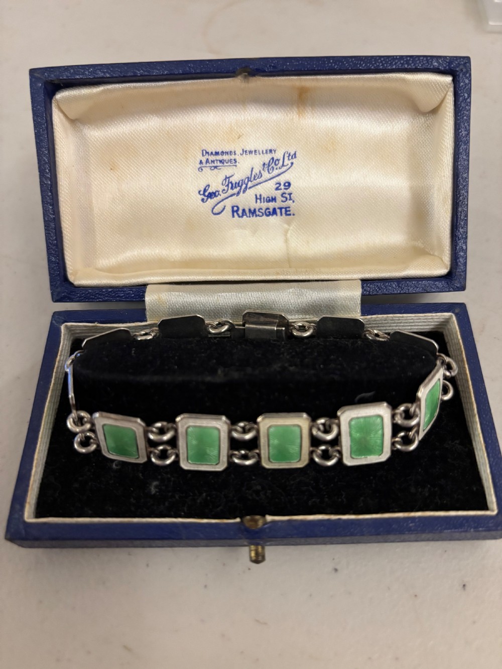 An Art Deco silver bracelet featuring rectangular green guilloche enamel panels resting inside an open fitted blue presentation box.