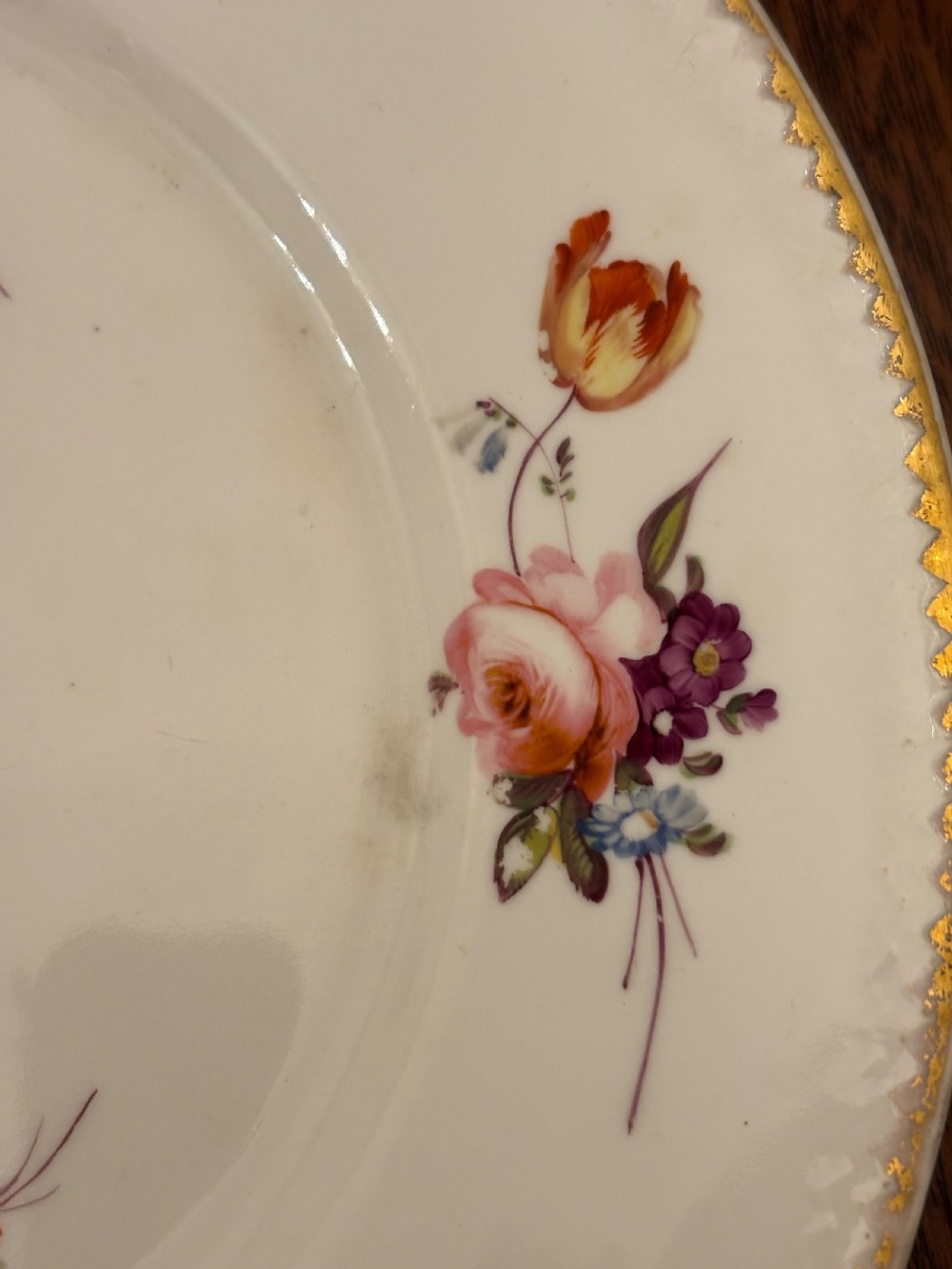 An antique off-white porcelain dessert service with hand-painted floral sprigs, scalloped edges, and gilded rims arranged against a black backdrop. View XV