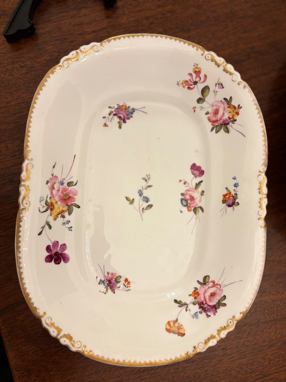 An antique off-white porcelain dessert service with hand-painted floral sprigs, scalloped edges, and gilded rims arranged against a black backdrop. View XXIV