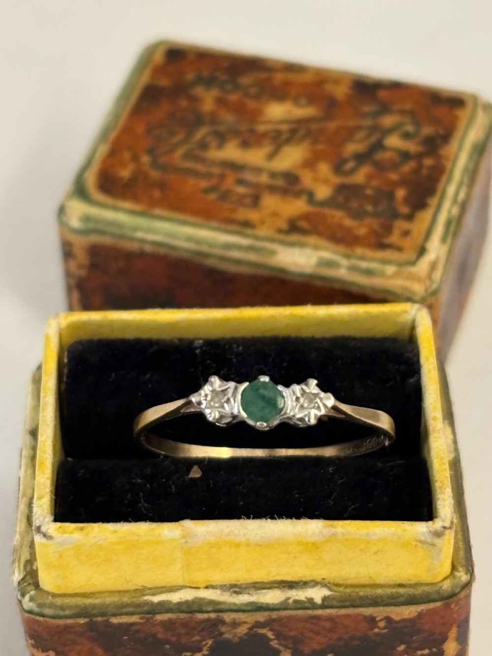 An antique gold ring featuring a central round green emerald flanked by two small diamonds set in star-shaped carved shoulders, placed inside a vintage velvet box.
