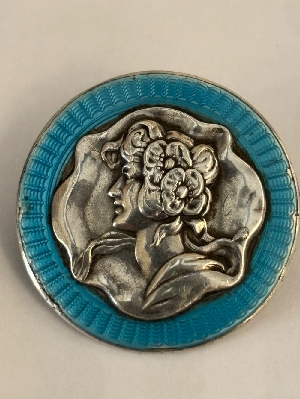 A circular silver brooch featuring a relief profile portrait of a woman framed by a bright turquoise blue guilloche enamel border.